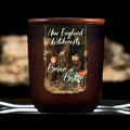 Image depicts a frosted amber candle jar with lid and a label depicting an oil painting of a child conversing with an old baker woman.