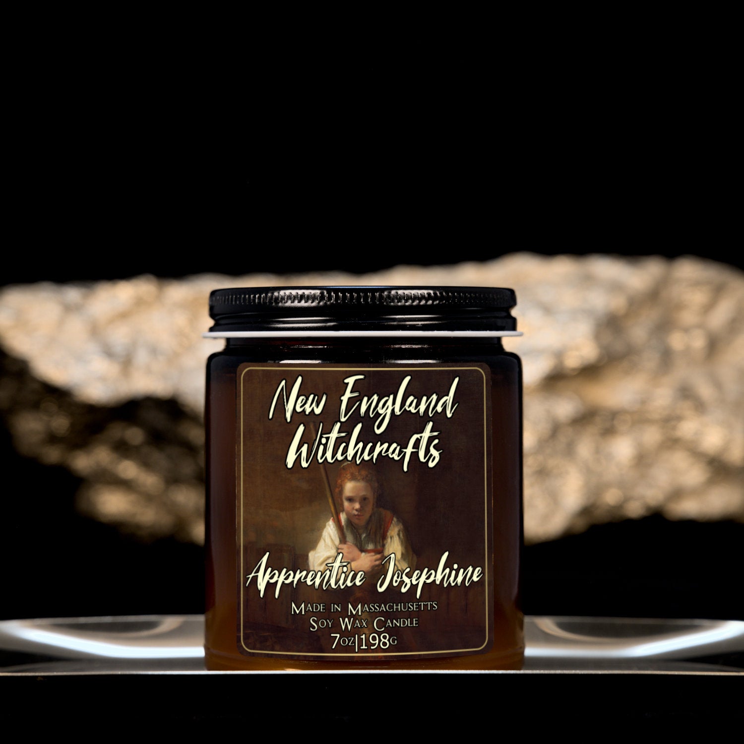 Image depicts an amber candle with black lid, smaller, and a label depicting an oil painting of a young witch apprentice holding her broom.