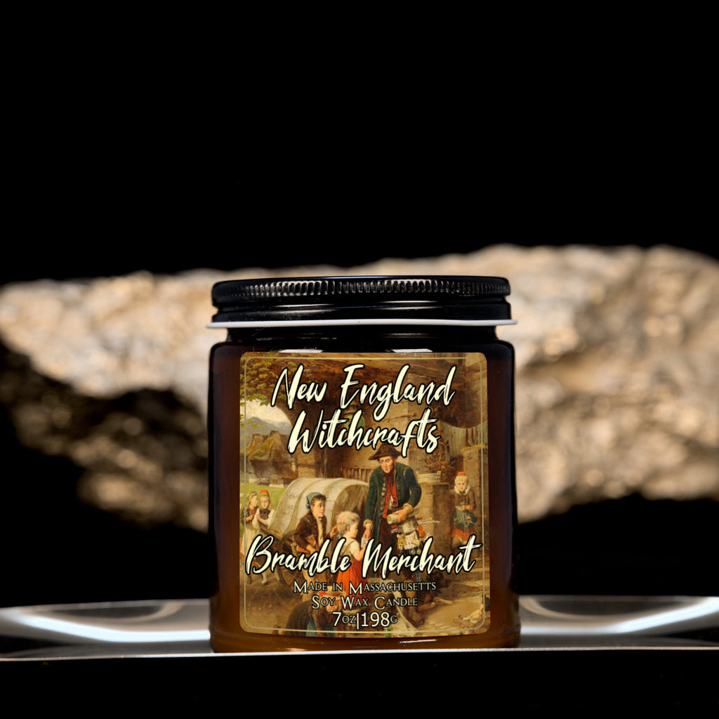 Image depicts an amber candle with black lid, smaller, and a label depicting an oil painting of a merchant arriving in town surrounded by curious children.
