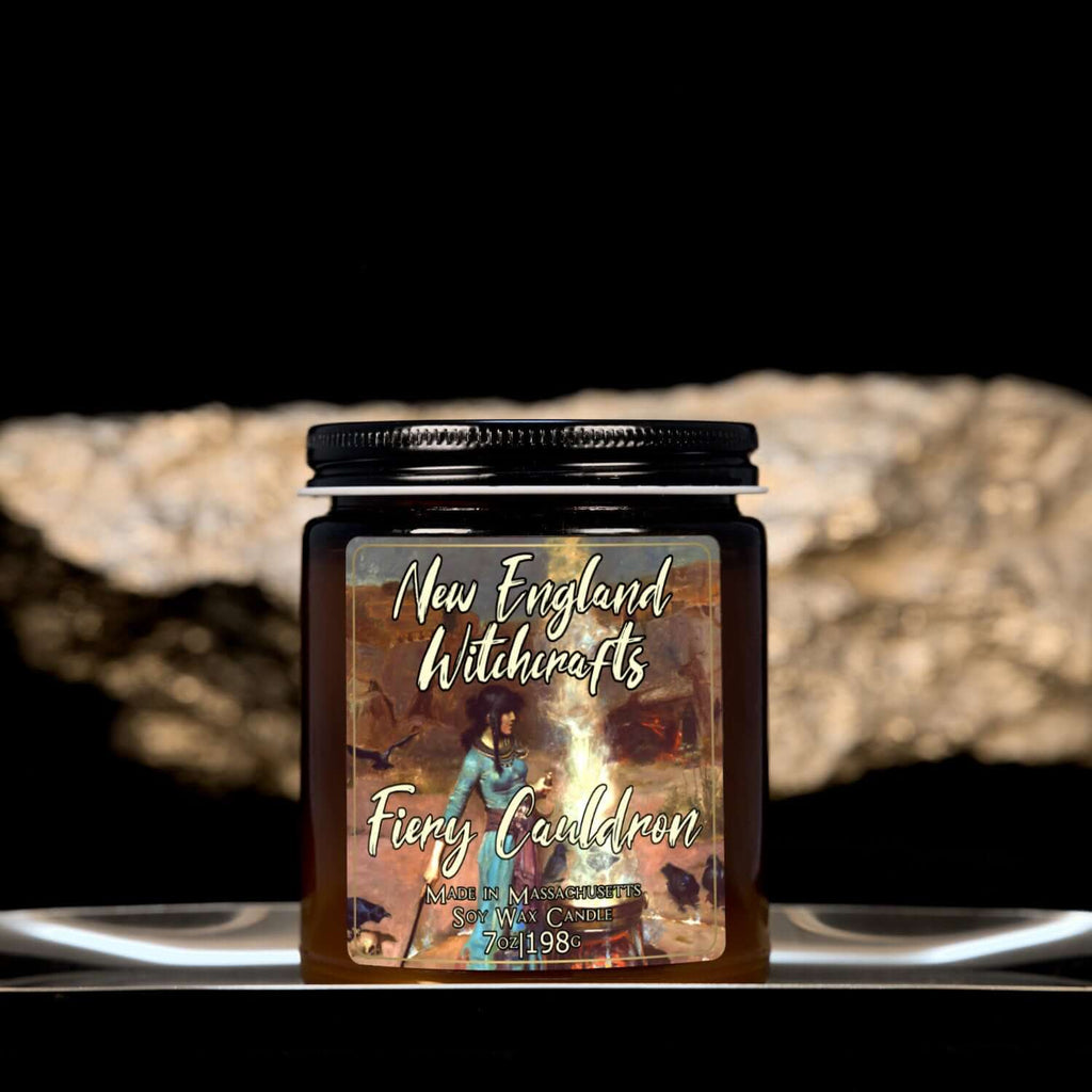 Image depicts an amber candle with black lid, smaller, and a label depicting an oil painting of a witch conjuring a spell over a fiery cauldron.