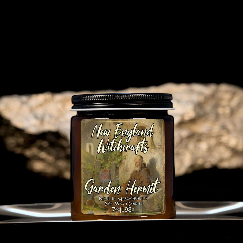 Image depicts an amber candle with black lid, smaller, and a label depicting an oil painting of a hermit sleeping against his shed.