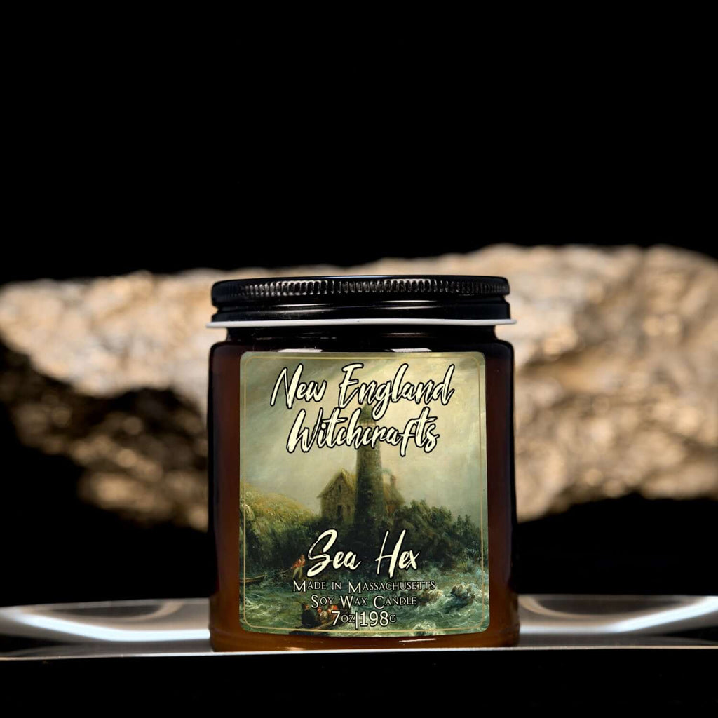 Image depicts an amber candle with black lid, smaller, and a label depicting an oil painting of stormy seas and a lighthouse.